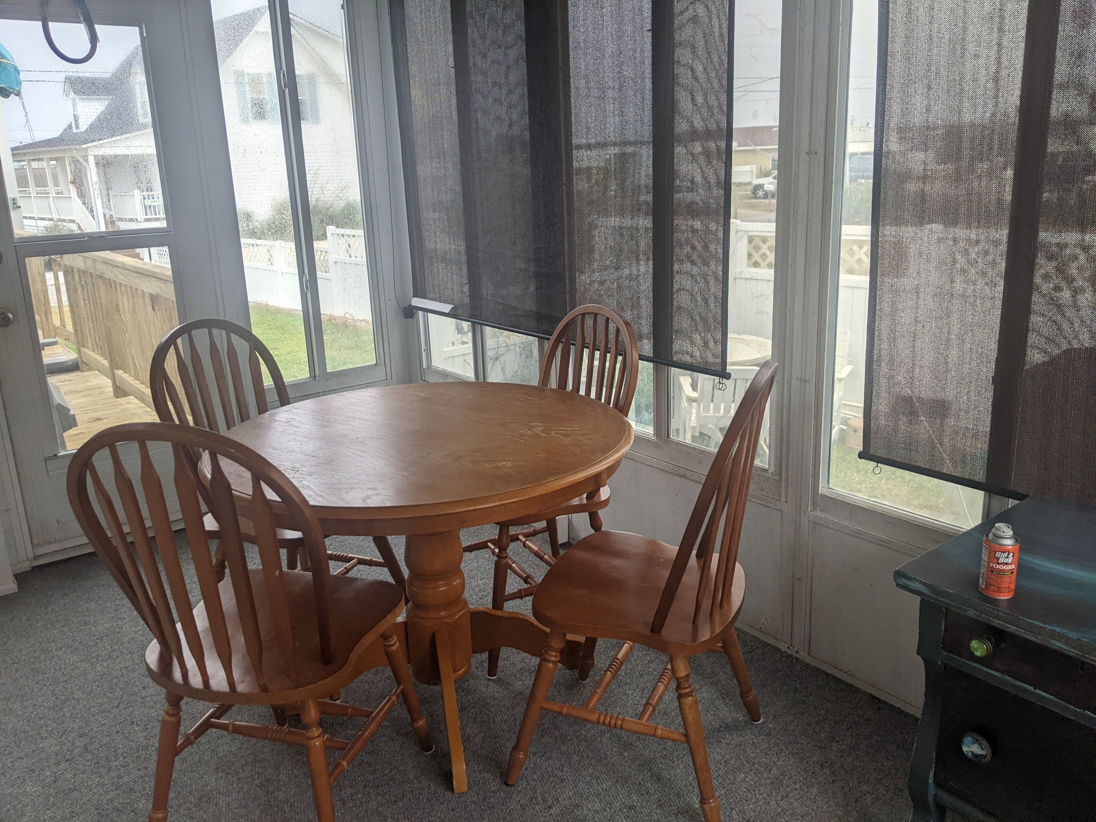 Une pièce avec une table ronde en bois et quatre chaises, de grandes fenêtres équipées de moustiquaires et une vue sur un jardin.