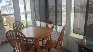 Une pièce avec une table ronde en bois et quatre chaises, de grandes fenêtres équipées de moustiquaires et une vue sur un jardin.