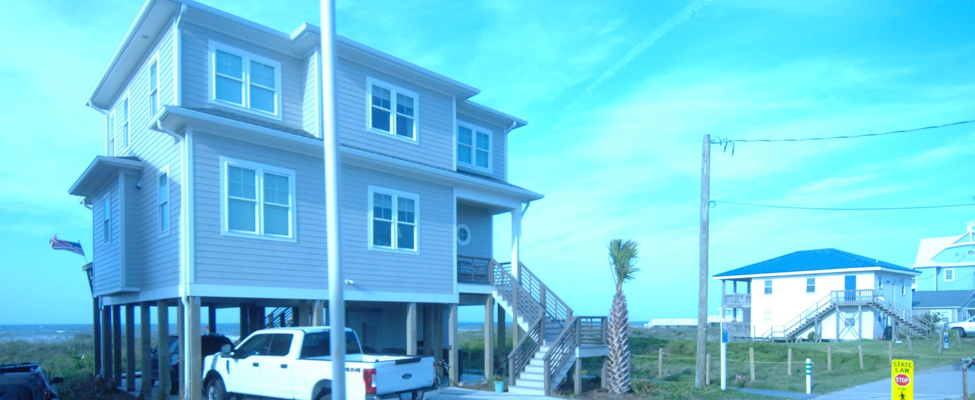 A beachfront property with a white truck parked in front, a staircase leading to a balcony, and a sign indicating public beach access.