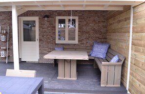 Eine überdachte Terrasse mit Holztisch und -bank, einer Tür und einem Fenster mit einer Chandelier.