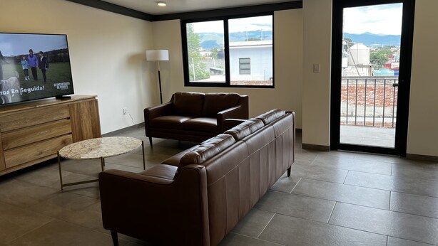 A spacious living room with a brown leather sofa, a small round table, and a television on a wooden cabinet.