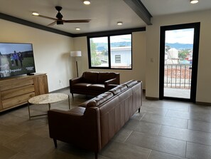A modern living room with a leather sofa, a television, a ceiling fan, and a view through a large window.