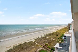 Une vue sur la plage, avec un ciel dégagé, une plage de sable et un bâtiment doté d’un balcon donnant sur la plage.