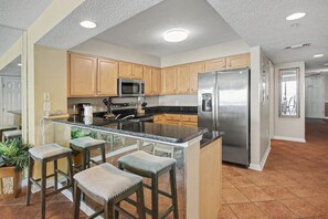 A kitchen with wooden cabinets, a stainless steel refrigerator, a microwave, and a bar area with stools.