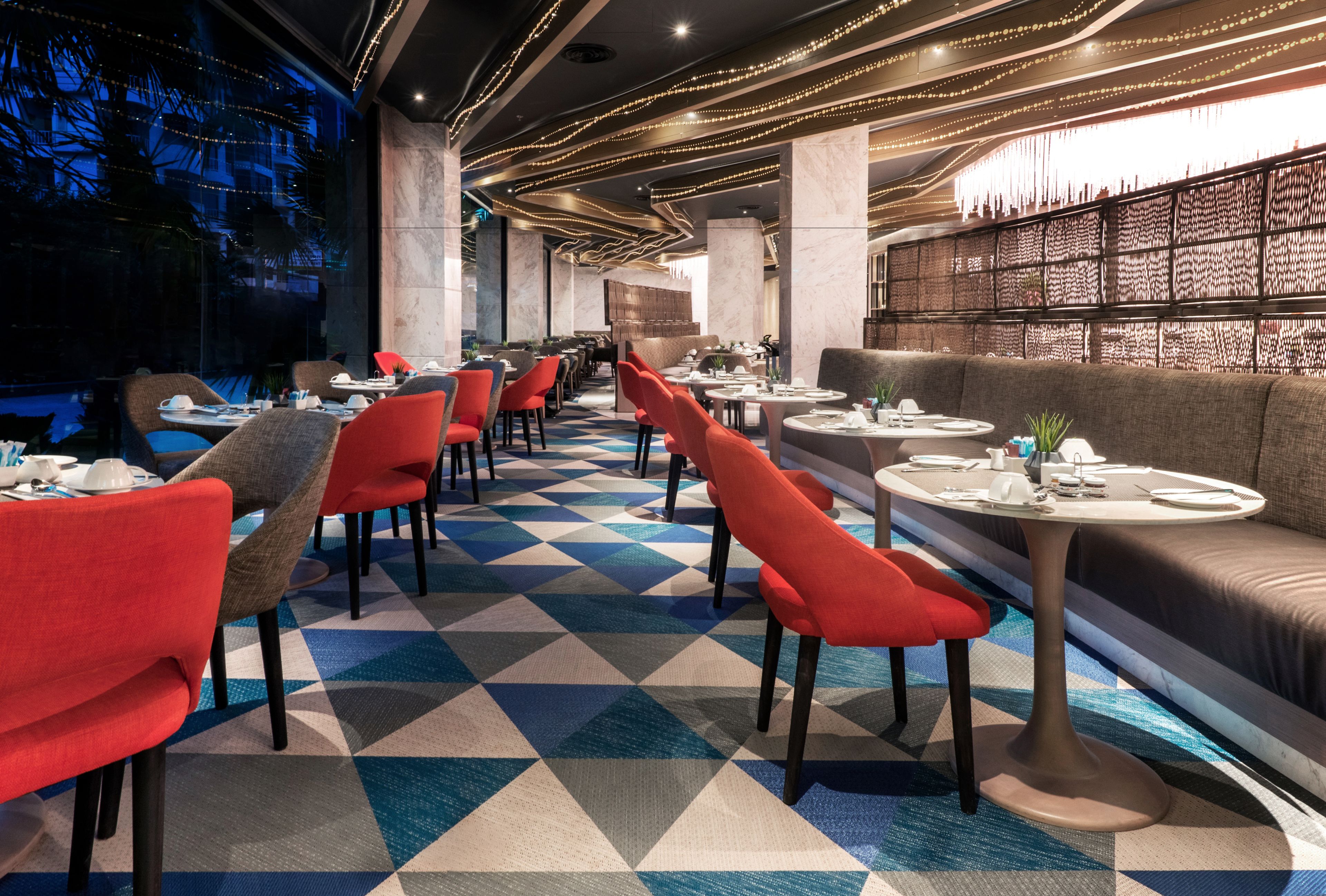 A modern restaurant interior with a geometric patterned floor, red and grey upholstered chairs, round tables, and a bar area with a patterned wall.