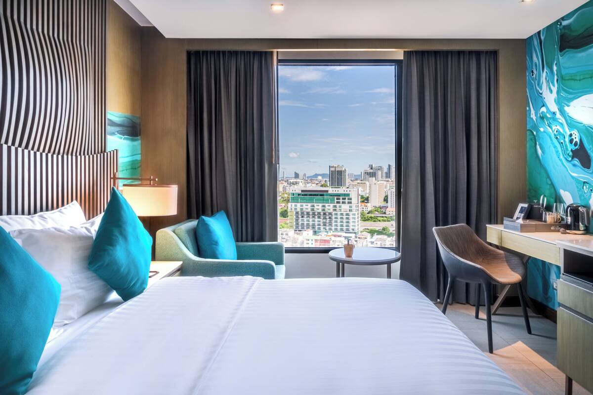 A modern hotel room with a large bed, a desk, a chair, and a view of the city.