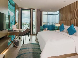 A modern hotel room with a large bed, a desk with a chair, a television, and a view of the city through large windows. - Mytt Hotel Pattaya (Pattaya)
