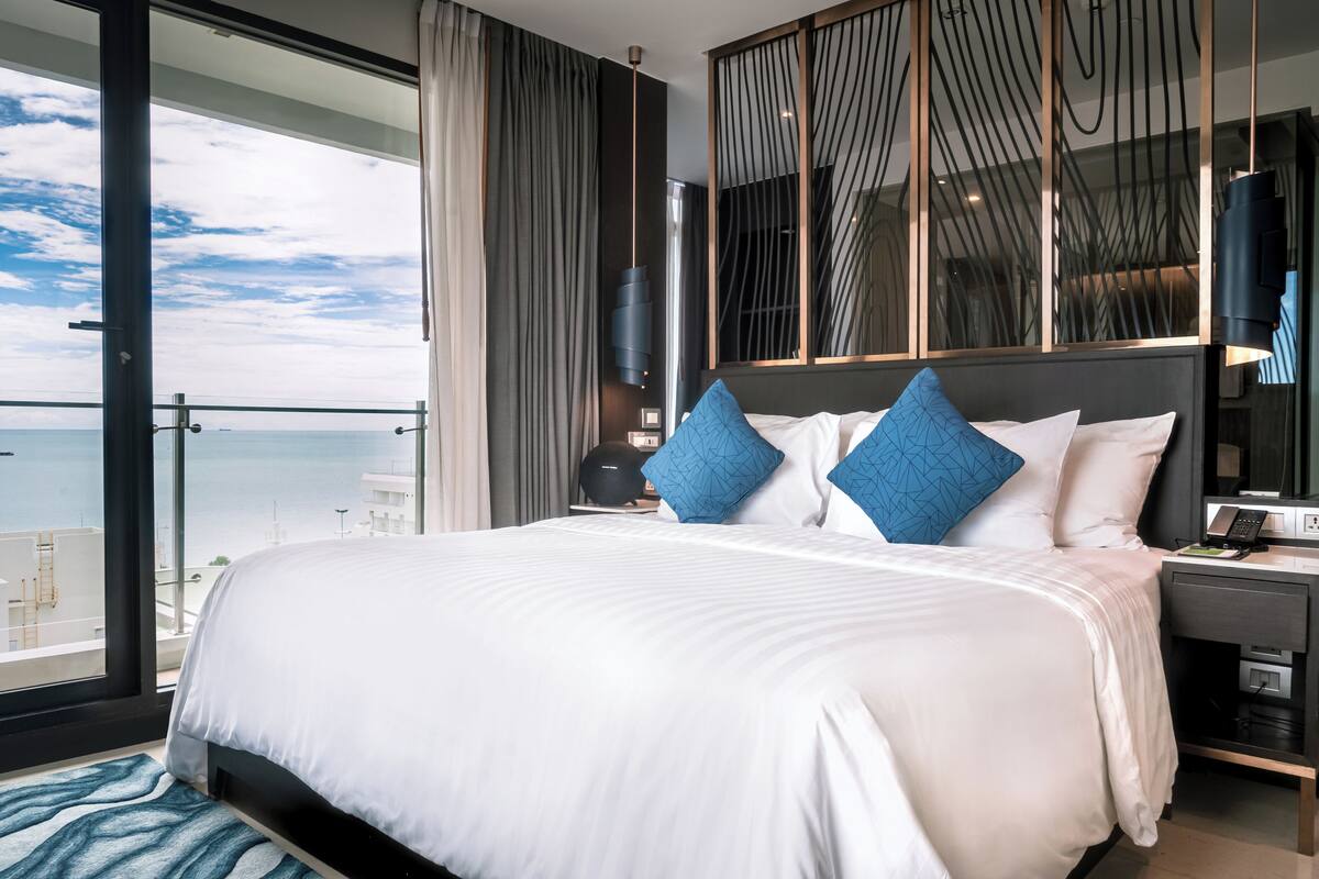 A modern hotel room with a large bed, a nightstand, a desk, and a view of the ocean.