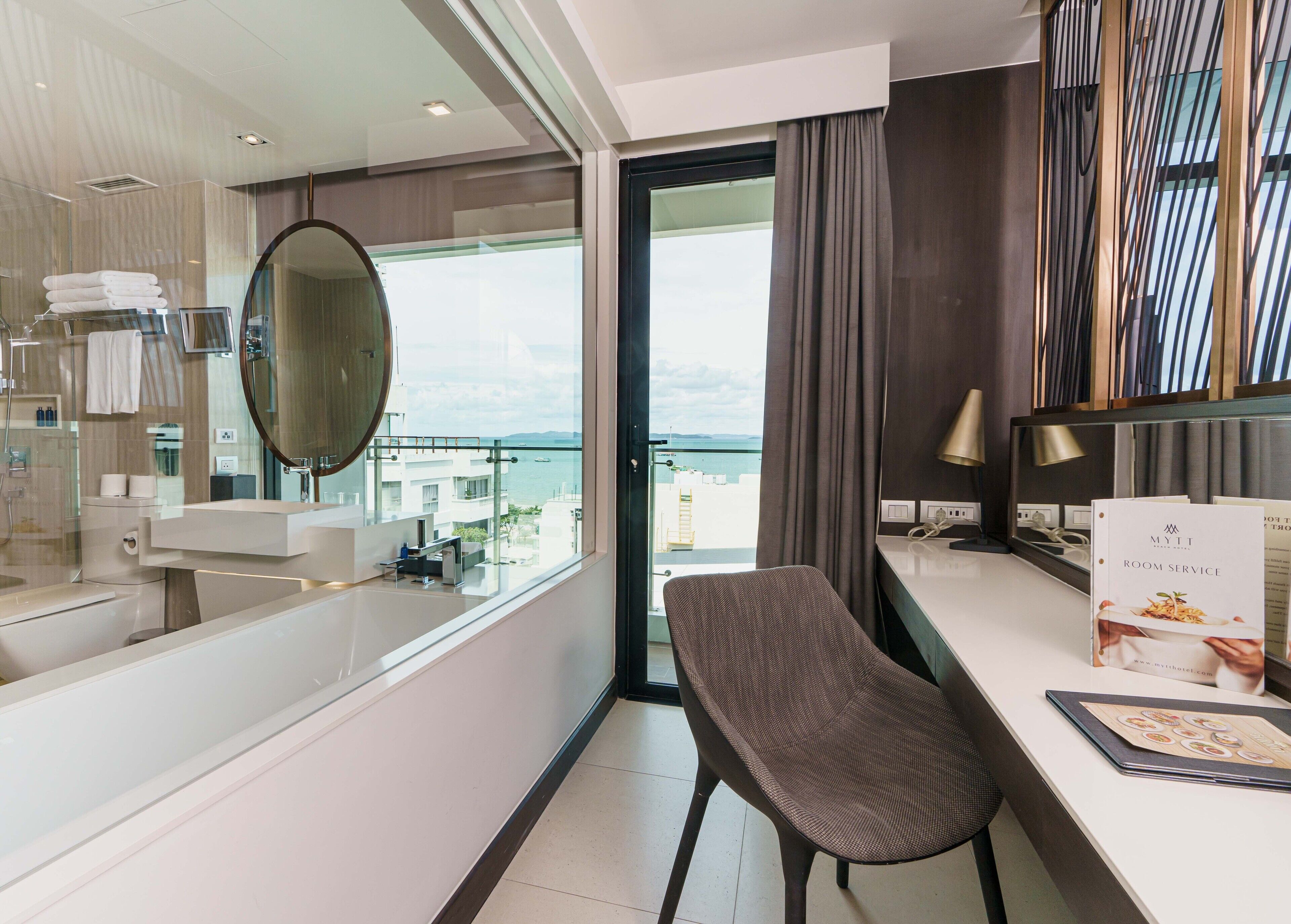 A modern hotel room with a balcony, a round mirror, a desk with a lamp, and a view of the sea.