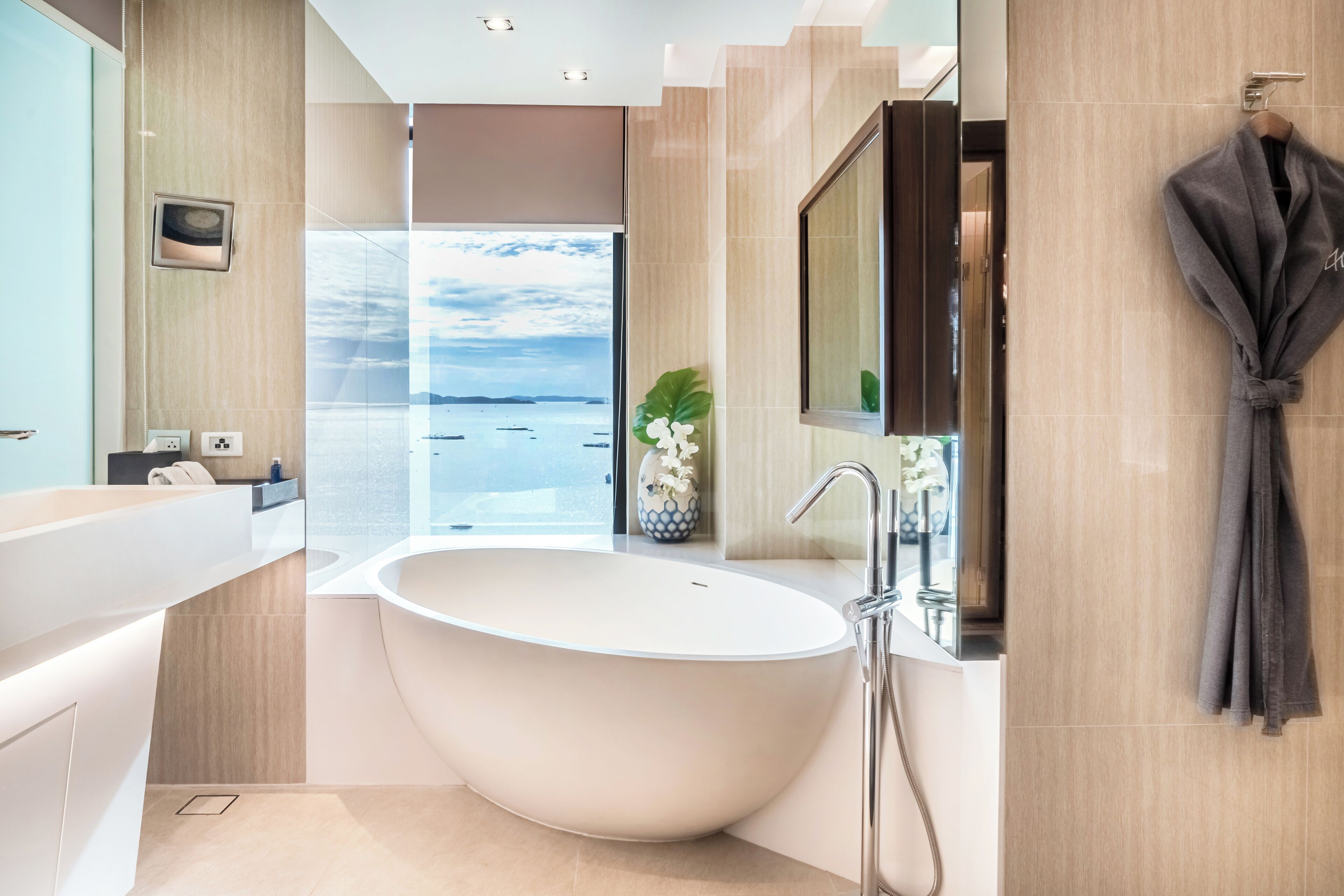 A modern bathroom with a freestanding bathtub, a large mirror, and a view of the sea.