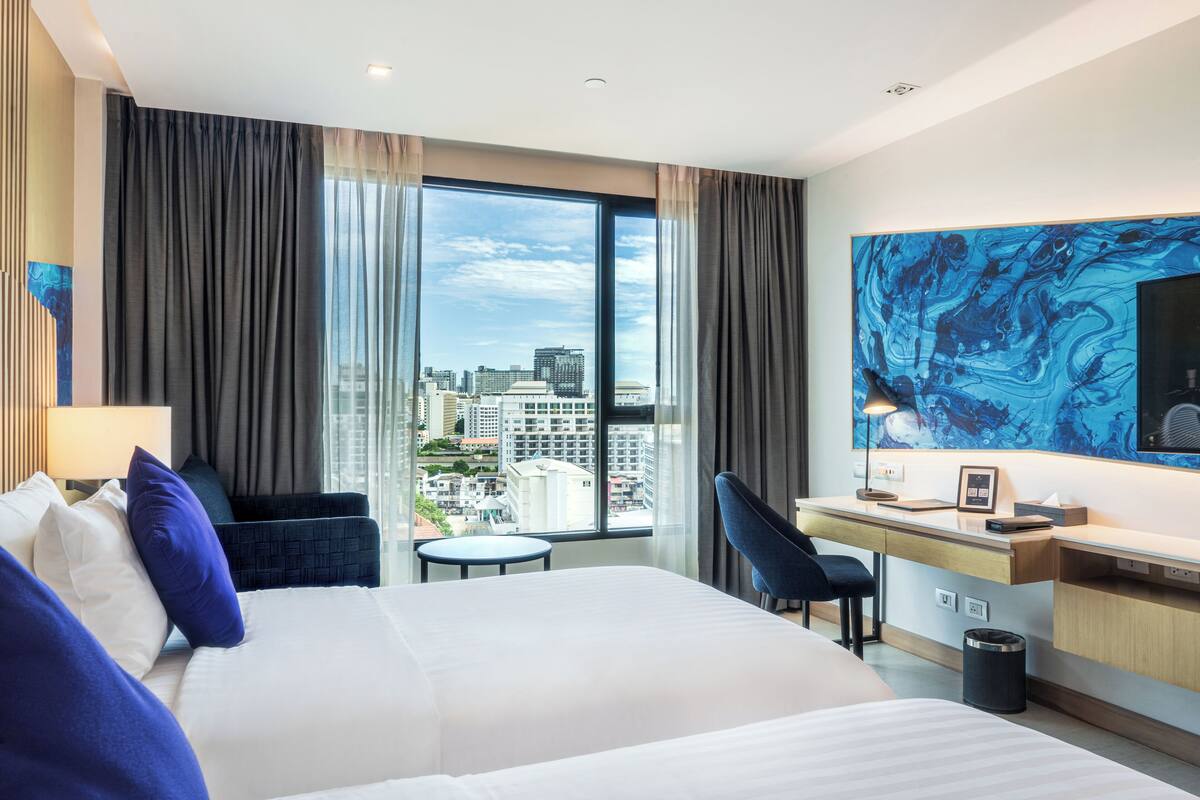 A hotel room with a large bed, a desk, a chair, and a view of the cityscape through large windows.