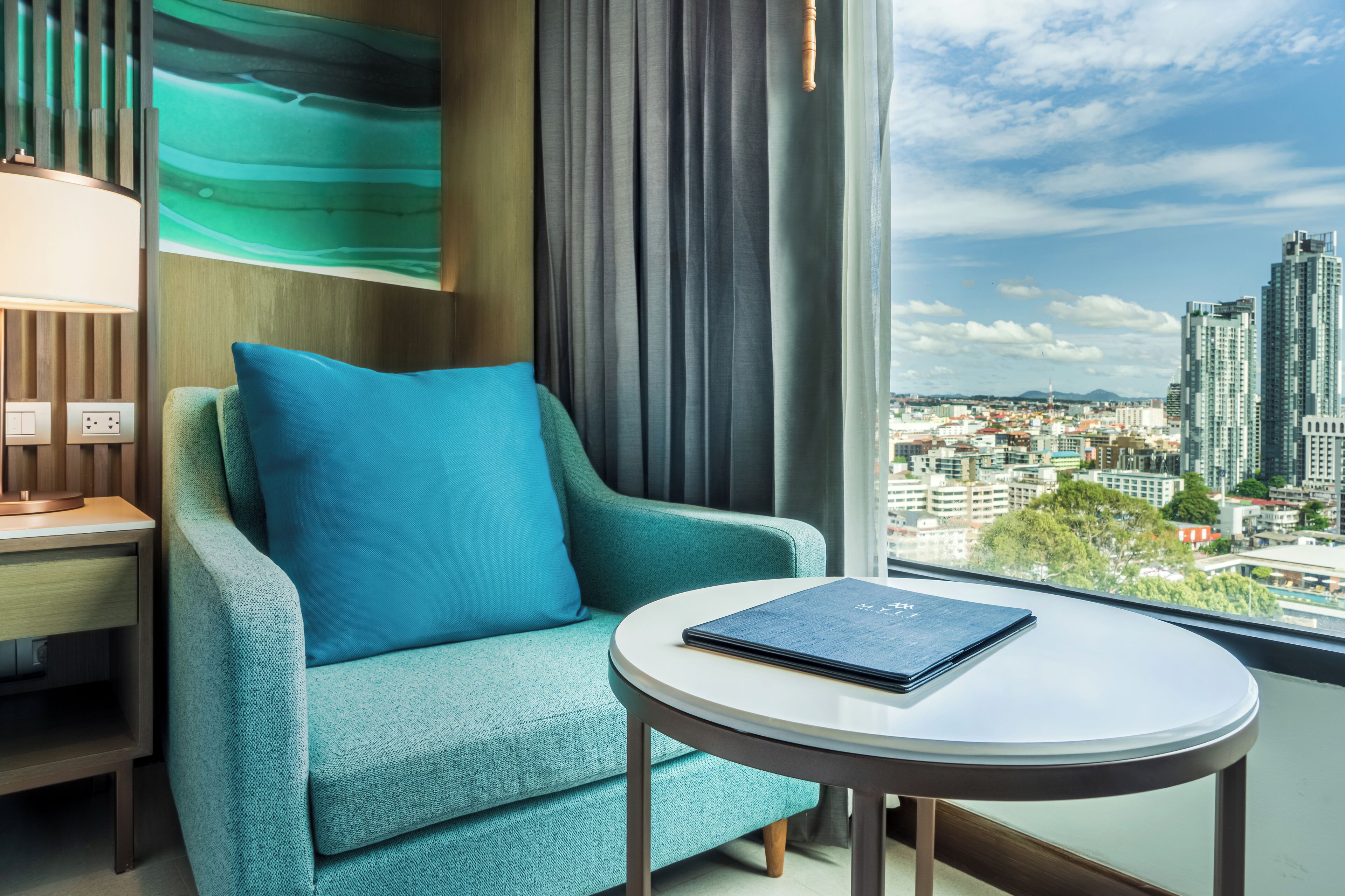 A teal armchair with a blue pillow, a round table with a book, and a city view through a large window.