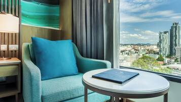 A teal armchair with a blue pillow, a round table with a book, and a city view through a large window.