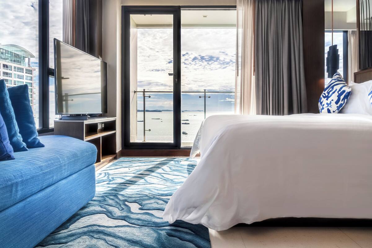 A hotel room with a large bed, a TV, and a view of the sea through a glass door.