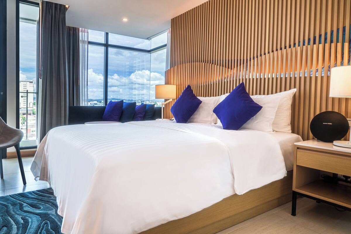 A hotel room with a large bed, a nightstand, a lamp, and a view of the city through large windows.