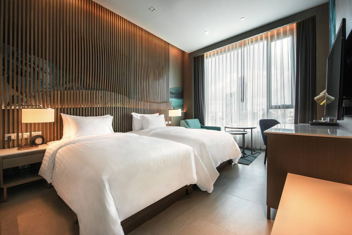 A modern hotel room with two beds, a desk, a chair, a TV, and a large window with curtains.
