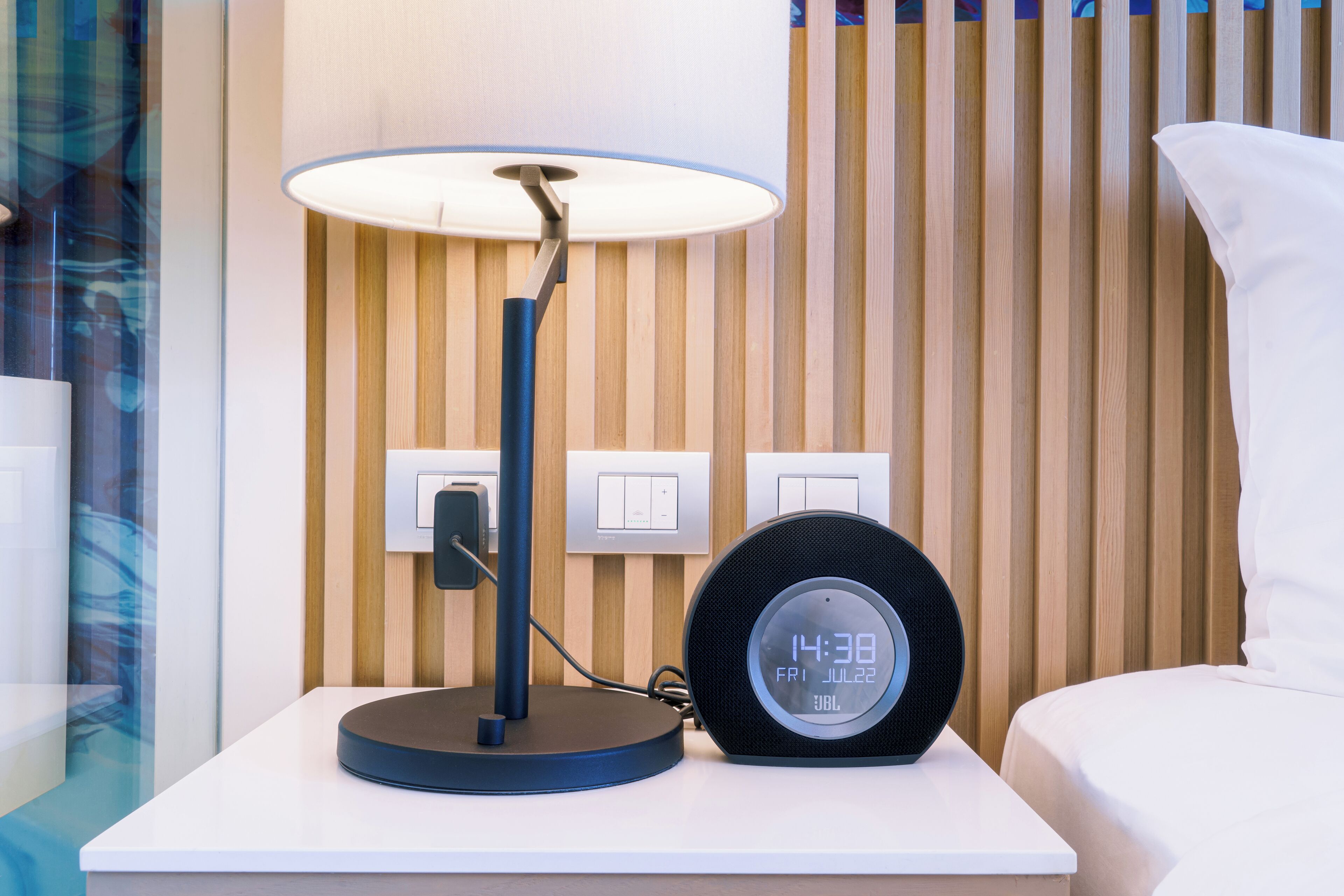A modern hotel room with a bedside table featuring a lamp, a digital clock, and a power outlet.
