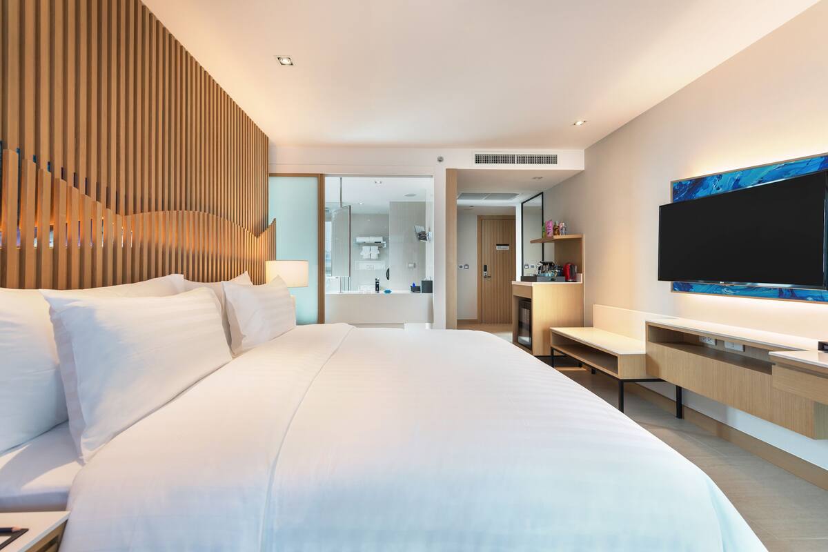 A modern hotel room with a large bed, a desk, a flat-screen TV, and a bathroom visible through an open door.