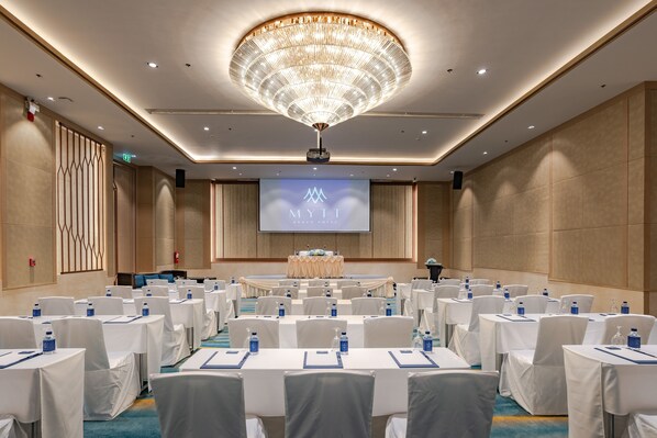 Un salón de conferencias con filas de mesas y sillas, una pantalla grande que muestra “MYTT BEACH HOTEL  ” y un candelabro.