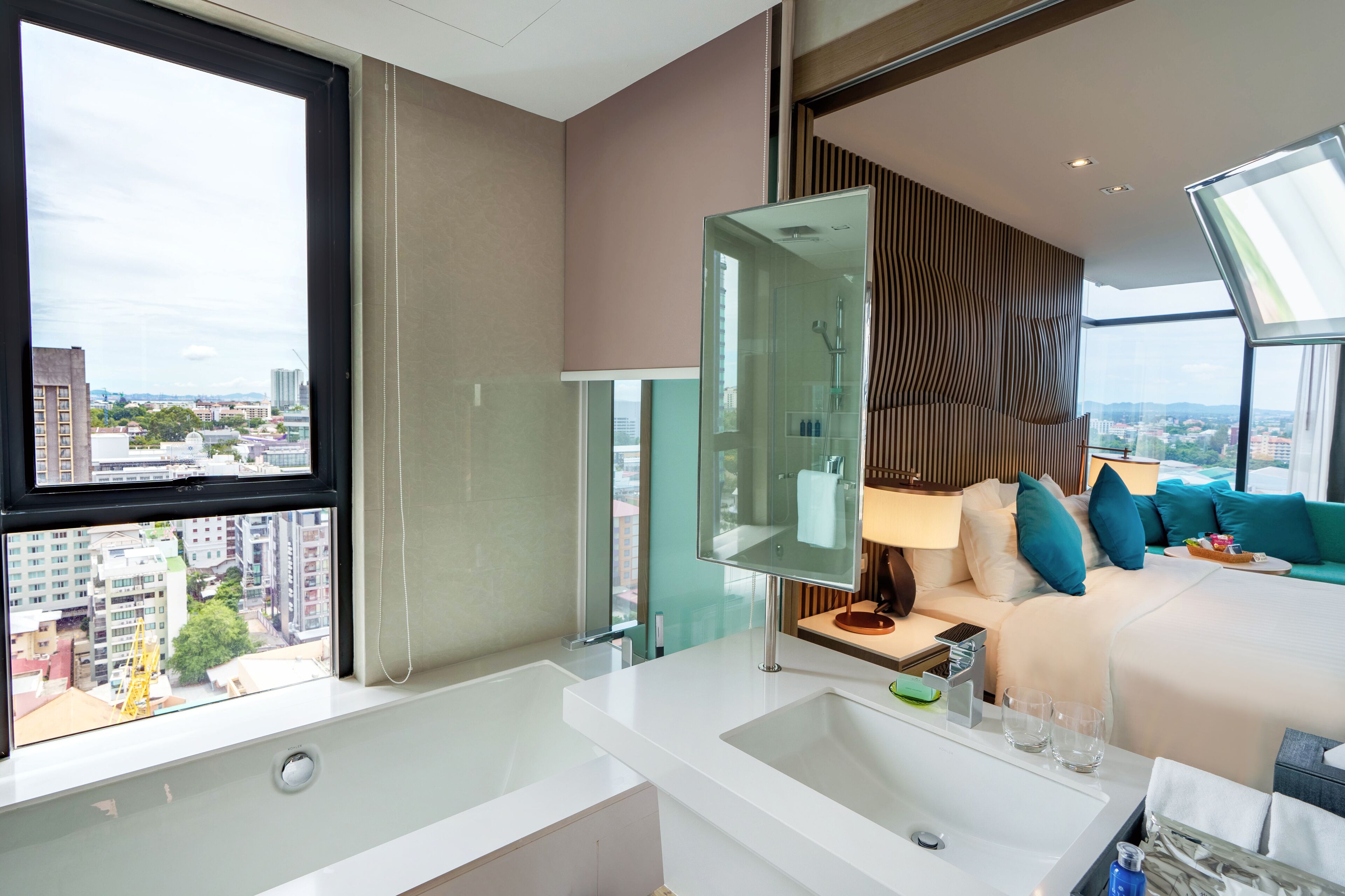 A modern hotel room with a large window, a bathtub, a vanity with a mirror, a bed with teal pillows, and a city view.