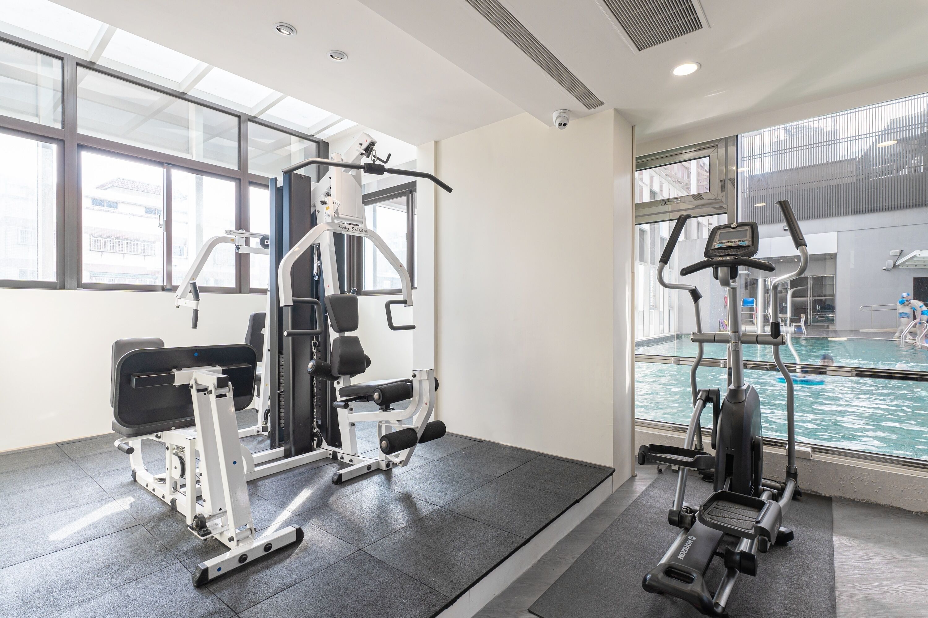 A gym with various exercise machines, a large window, and a pool visible through the glass.