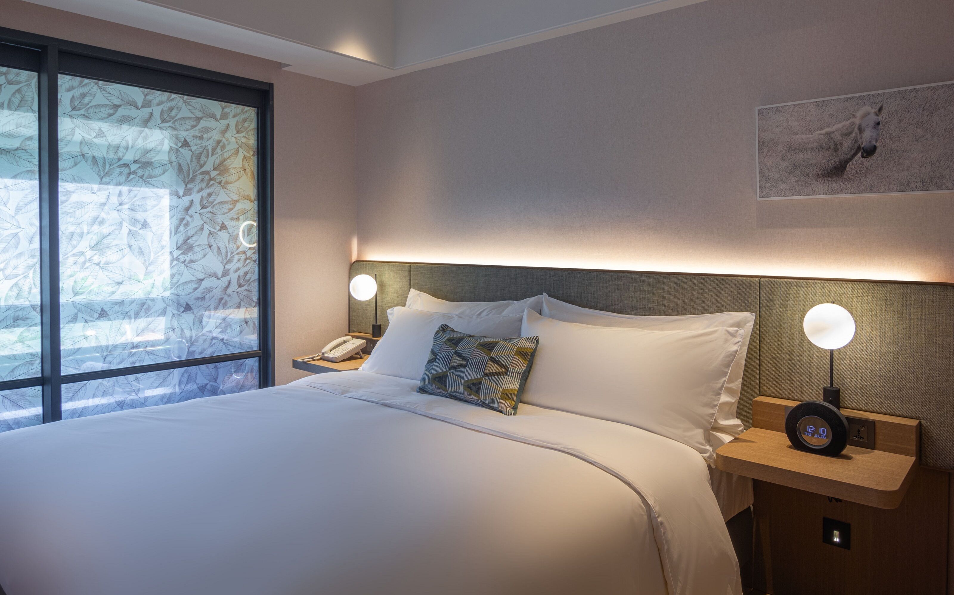 Ein Hotelzimmer mit einem großen Bett, einem Nachttisch mit Telefon, einer Wanduhr und einem Fenster mit Blattmuster.