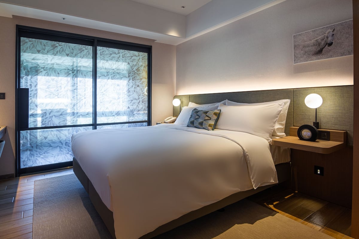 Ein modernes Hotelzimmer mit einem großen Bett, einem Nachttisch mit einer Uhr und einem großen Fenster mit mattiertem Glas.