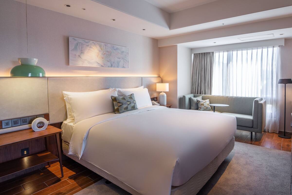 Ein modernes Hotelzimmer mit einem großen Bett, Nachttischen, einem Schreibtisch, einem Sessel und einem großen Fenster mit Vorhängen.