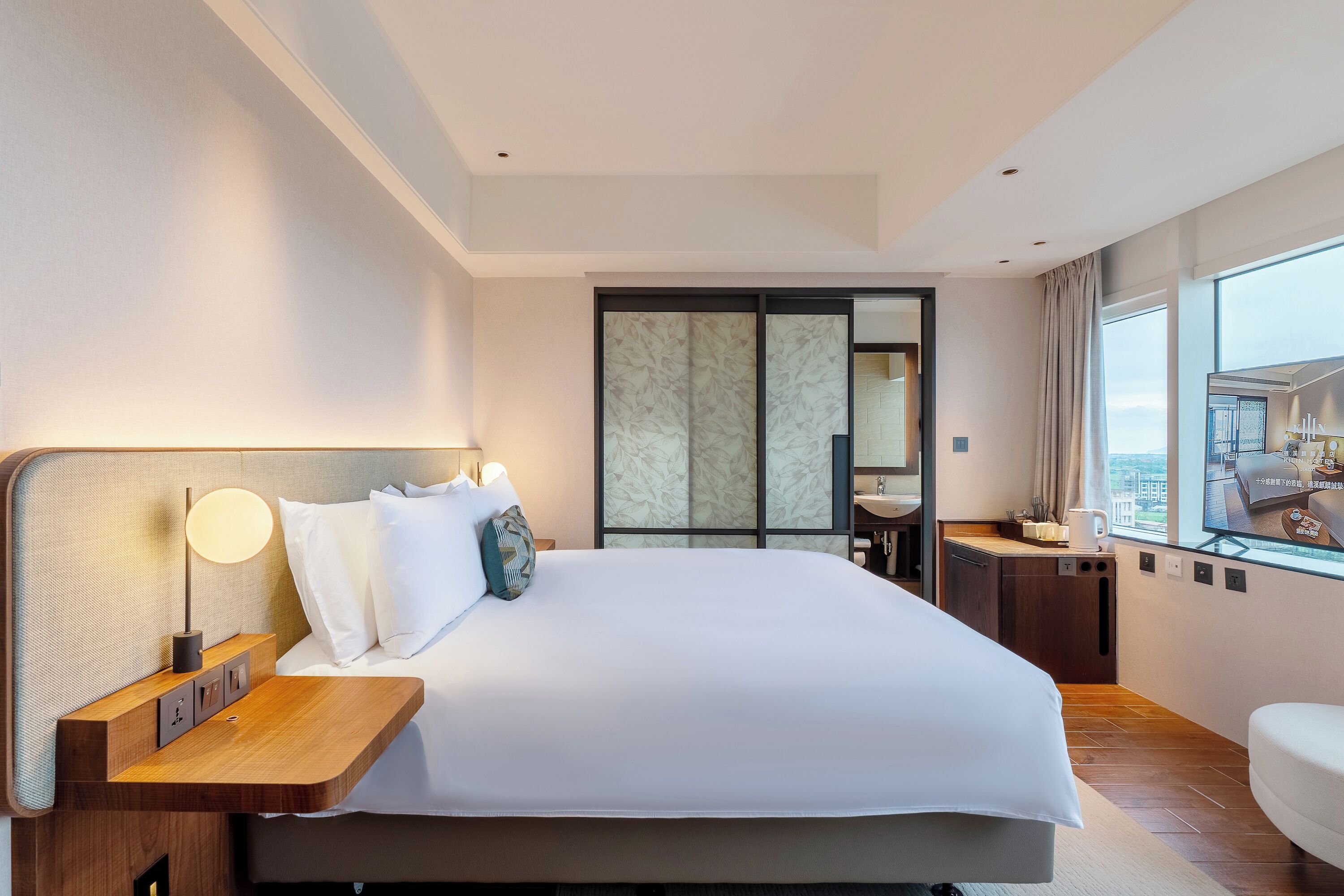 A modern hotel room with a large bed, bedside table, lamp, and a view of the city through a large window.