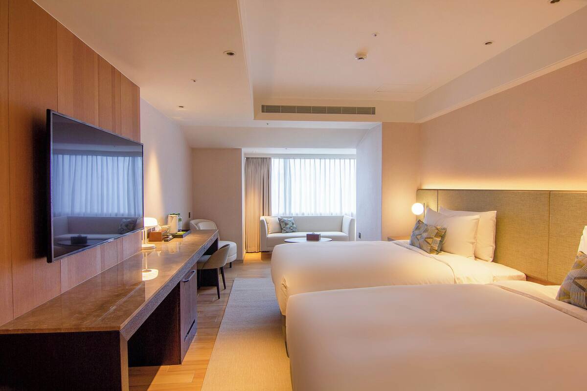 Ein Hotelzimmer mit Flachbildfernseher, Schreibtisch mit Stuhl, zwei Betten, einer Sitzecke und einem großen Fenster mit Vorhängen.