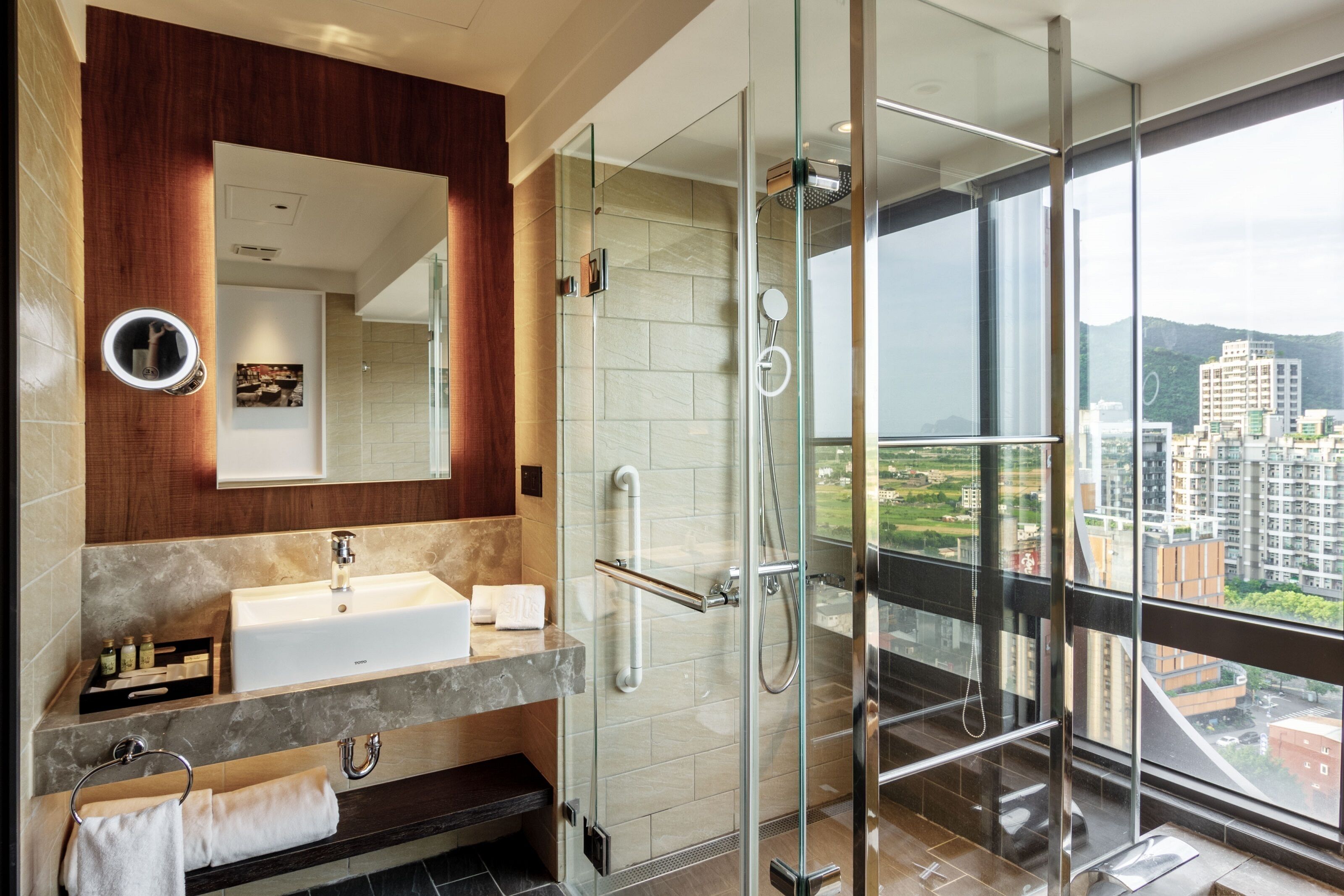 A modern bathroom with a glass-enclosed shower, a large mirror, and a view of a cityscape through large windows.