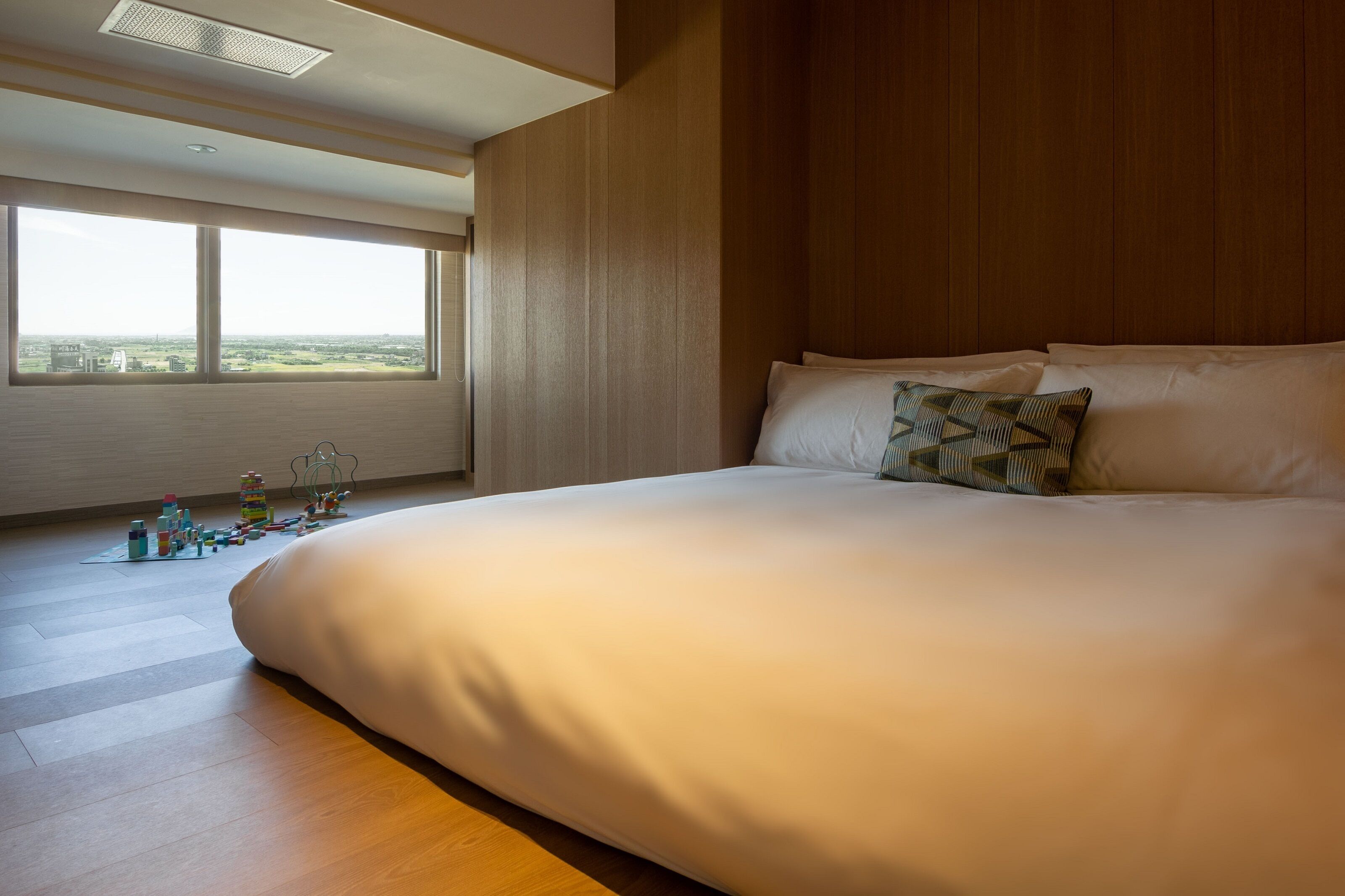 A large bed with white linens and a single patterned pillow, a wooden floor, and a large window with a view of the city.