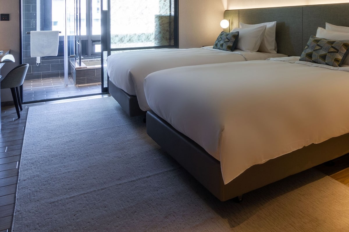 Ein Hotelzimmer mit zwei Betten, einem Schreibtisch, einem Stuhl und einem kleinen Tisch mit einer Lampe.