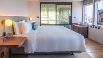 A modern hotel room with a large bed, a bedside table with a lamp, a small desk with a chair, and a window with a city view.