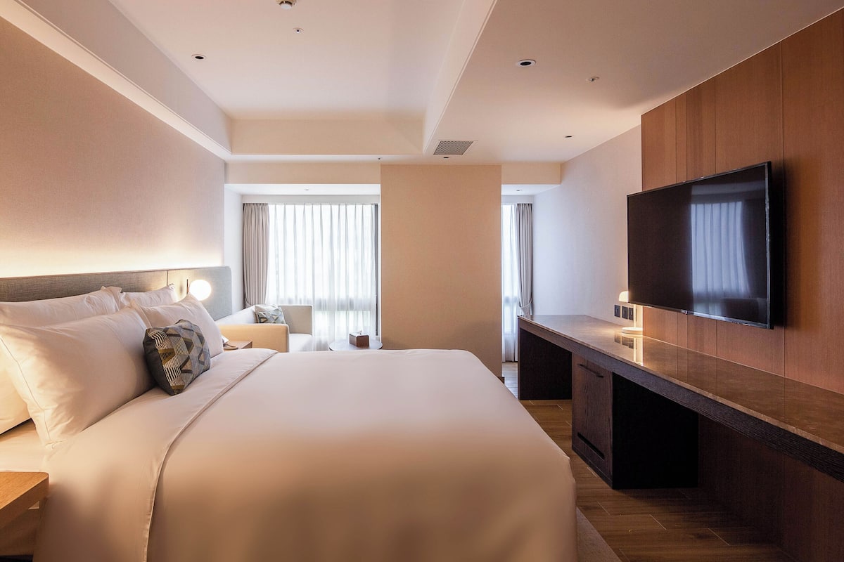 Ein modernes Hotelzimmer mit einem großen Bett, einem flachbildfernseher an der Wand, einem Schreibtisch und einem Fenster mit Vorhängen.