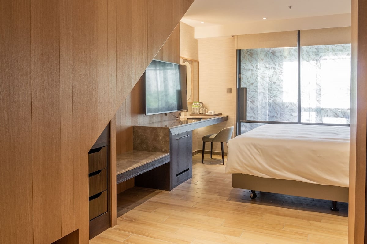 Ein modernes Hotelzimmer mit einer Holztreppe, einem Schreibtisch mit Flachbildfernseher, einem Bett und großen Fenstern.