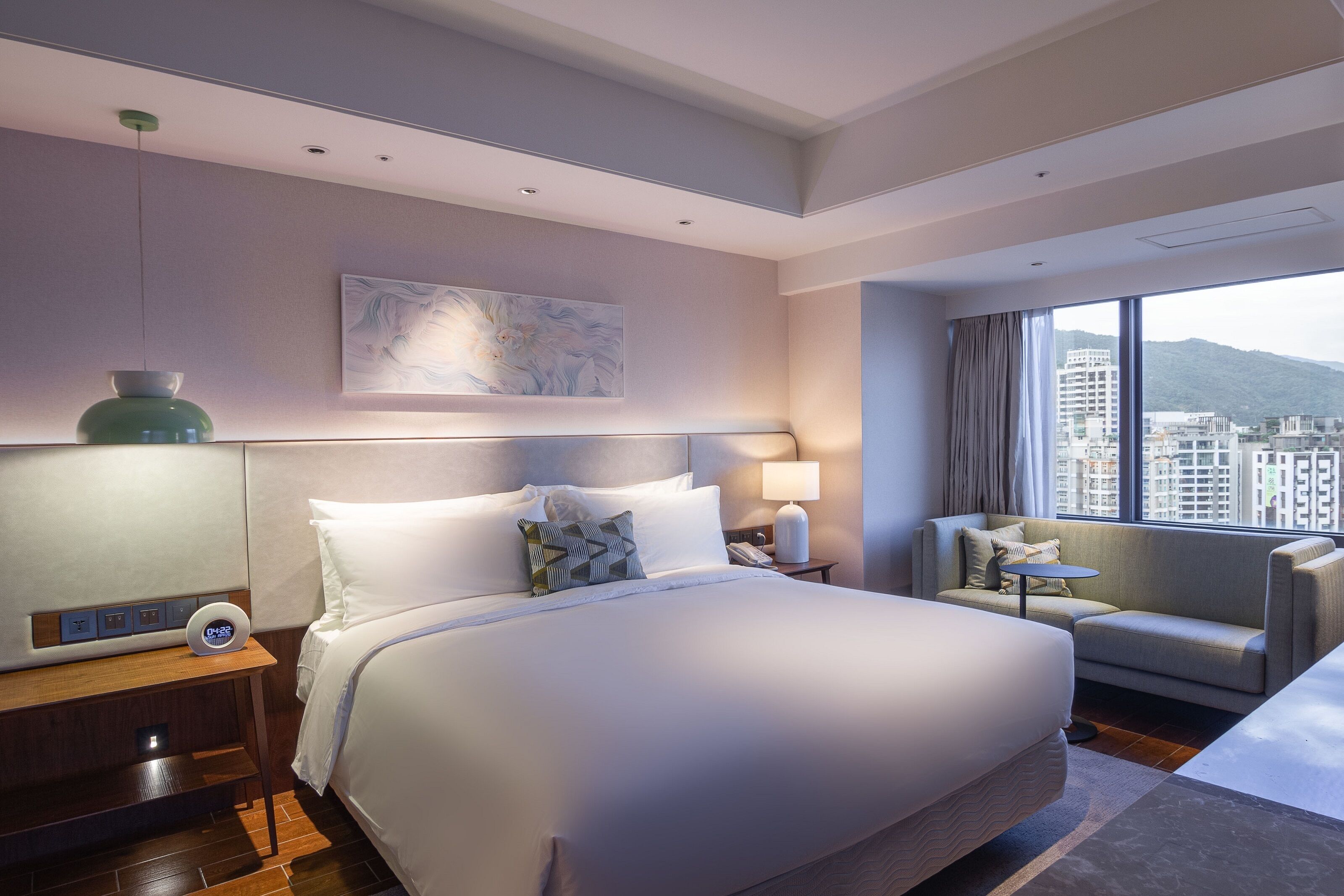 Una habitación de hotel moderna con una cama grande, una mesita de noche con un reloj, un revistero y un sofá, con vistas a la ciudad.