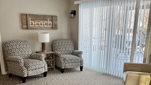 Ein Wohnzimmer mit einem gemusterten Sessel, einer Couch und einem kleinen Tisch mit einer Lampe. An der Wand hängt ein Schild mit der Aufschrift 'beach' und ein Fenster mit Jalousien.
