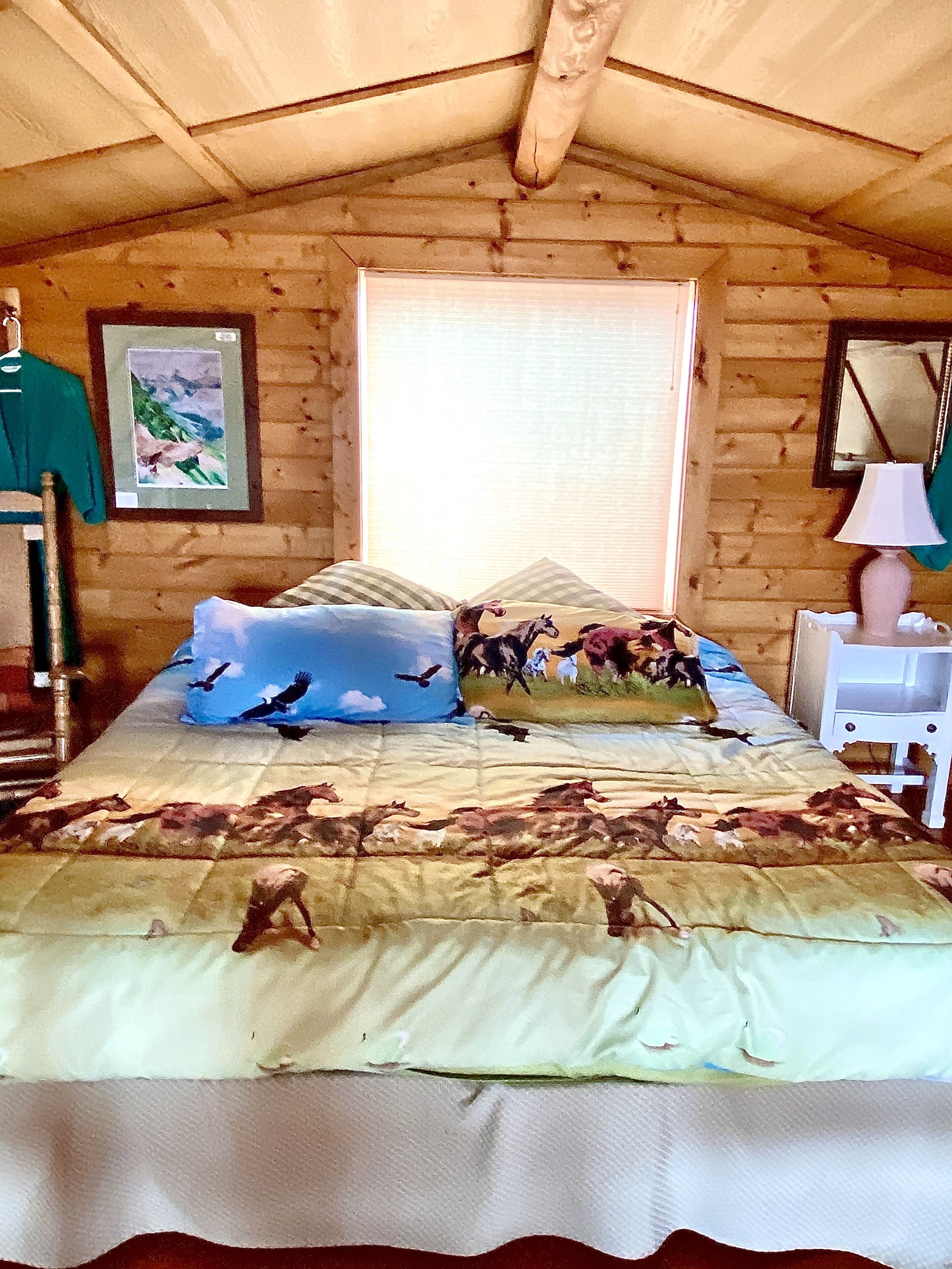 Een slaapkamer met een houten plafond, een bed met een beddekleed met dessin, een raam met zonwering, een schilderij aan de muur en een nachtkastje met een lamp.