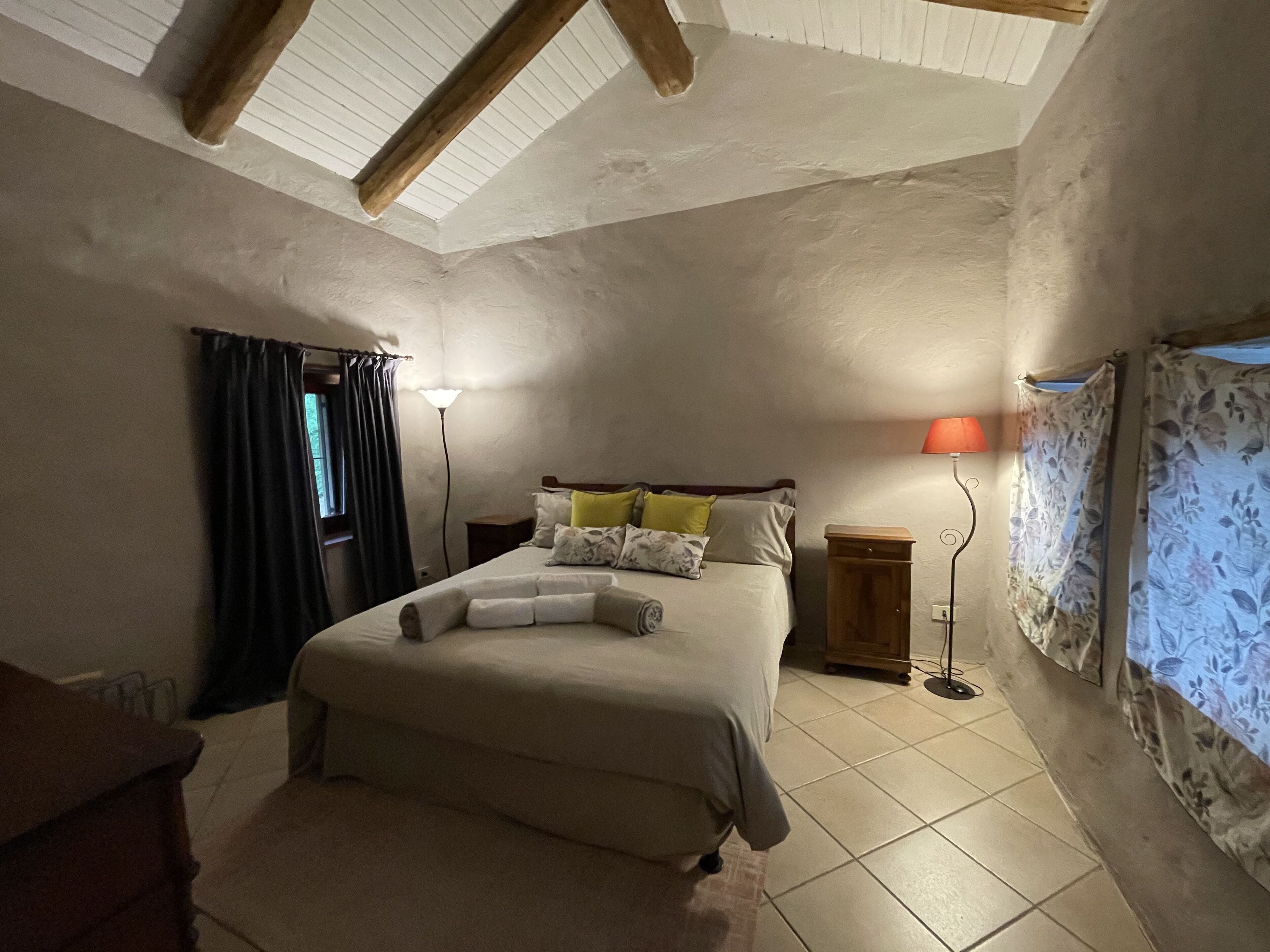 Une chambre à coucher comprenant un lit, une table de chevet, une lampe, une fenêtre avec des rideaux et une poutre en bois au plafond.