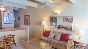 A cozy living room with a sofa, two chairs, a coffee table, and a small dining table. There is a ceiling fan, wall-mounted lamps, and framed artwork. - Direct Oceanfront Condo!  Sea Turtles & Beautiful Sunsets from the Lanai (Kailua-Kona)