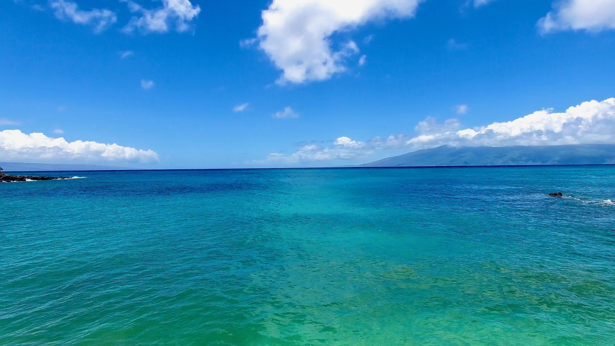 A clear view of turquoise ocean water with a distant mountain and a rocky shoreline.