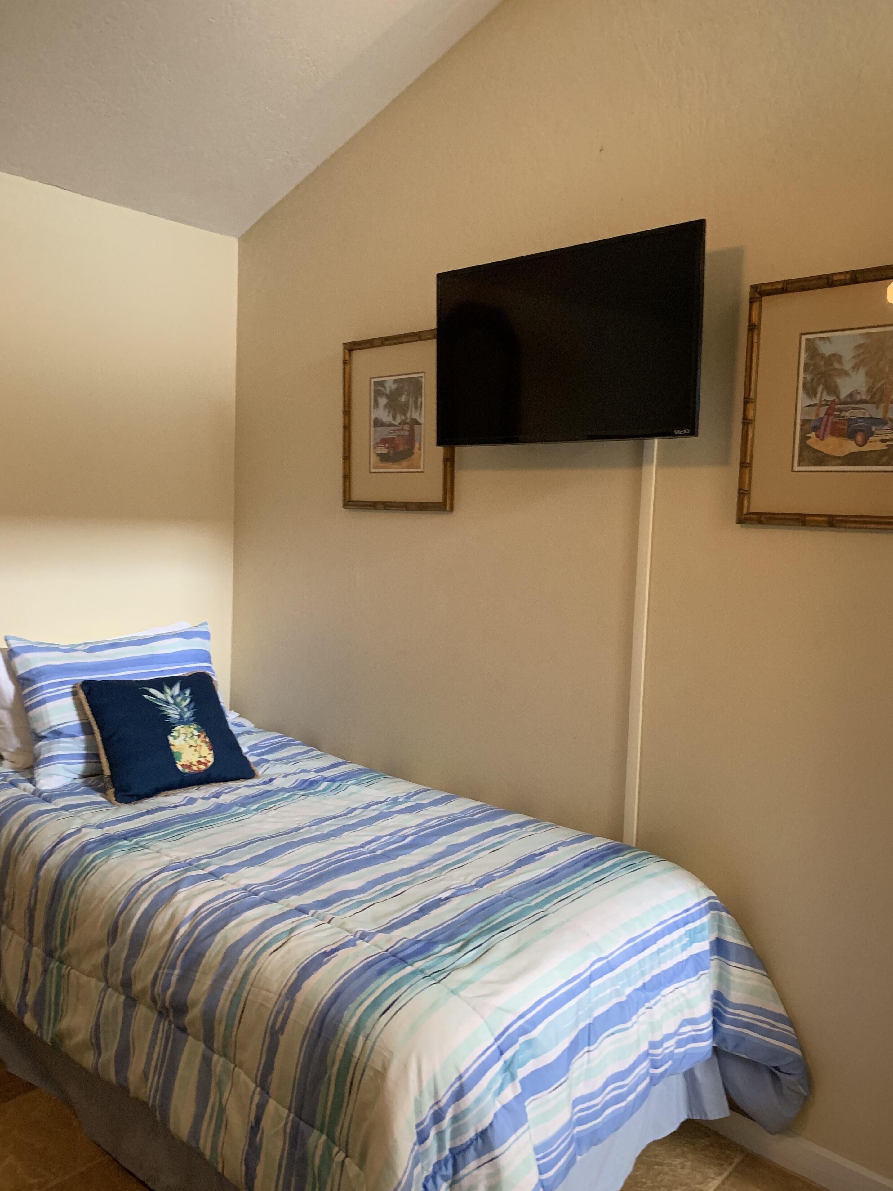 A single bed with a striped bedspread, a pineapple pillow, and a TV mounted on the wall.