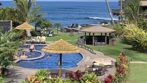 Un área de piscina con una pileta circular, sillas de descanso y sombrillas. Se observan palmeras y vistas al océano.