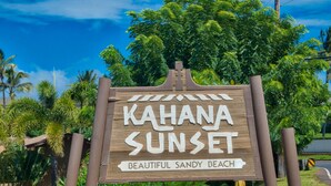 Un letrero de madera con el texto "KAHANA SUNSET" y "PLAYA DE ARENA HERMOSA", una flecha señalando a la izquierda hacia "OFICINA", rodeado de exuberante follaje y flores.