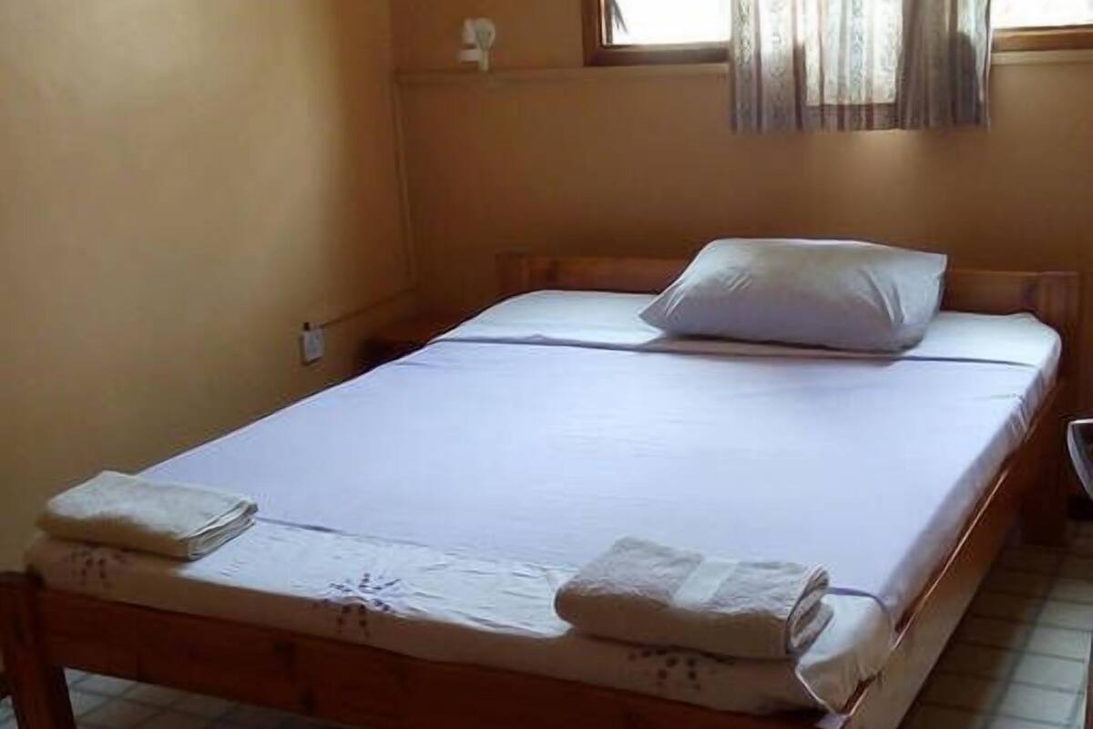 A single bed with white linens and a wooden frame in a room with tiled flooring and a window with curtains.