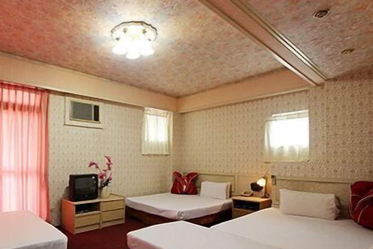 A hotel room with three single beds, a television on a wooden stand, and a small table with a lamp.