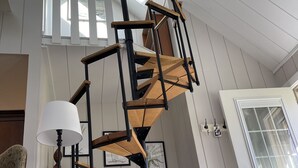Une pièce avec un escalier en colimaçon, une lampe, une chaise, une petite table et une porte avec une fenêtre.