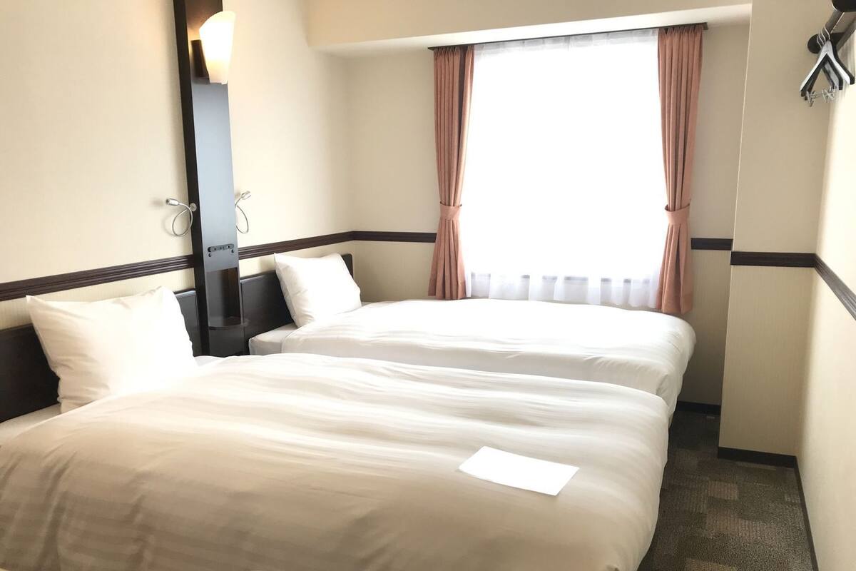 A hotel room with two beds, a nightstand, a mirror, a window with curtains, and a wall-mounted coat rack.