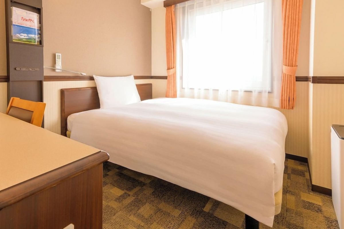A hotel room with a bed, a desk, a chair, a window with curtains, and a wall-mounted lamp.